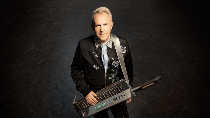 Howard Jones, dressed in a suit, stands with a keyboard in hand, looking ready to play