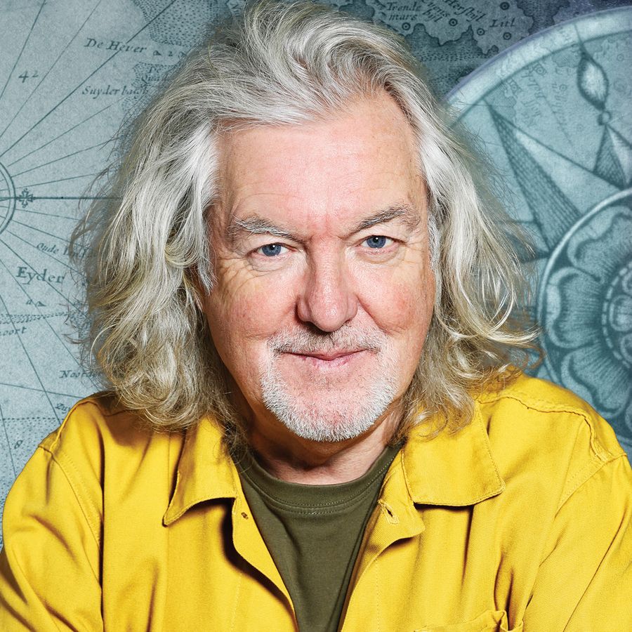 James May with long hair in a yellow shirt stands in front of a map