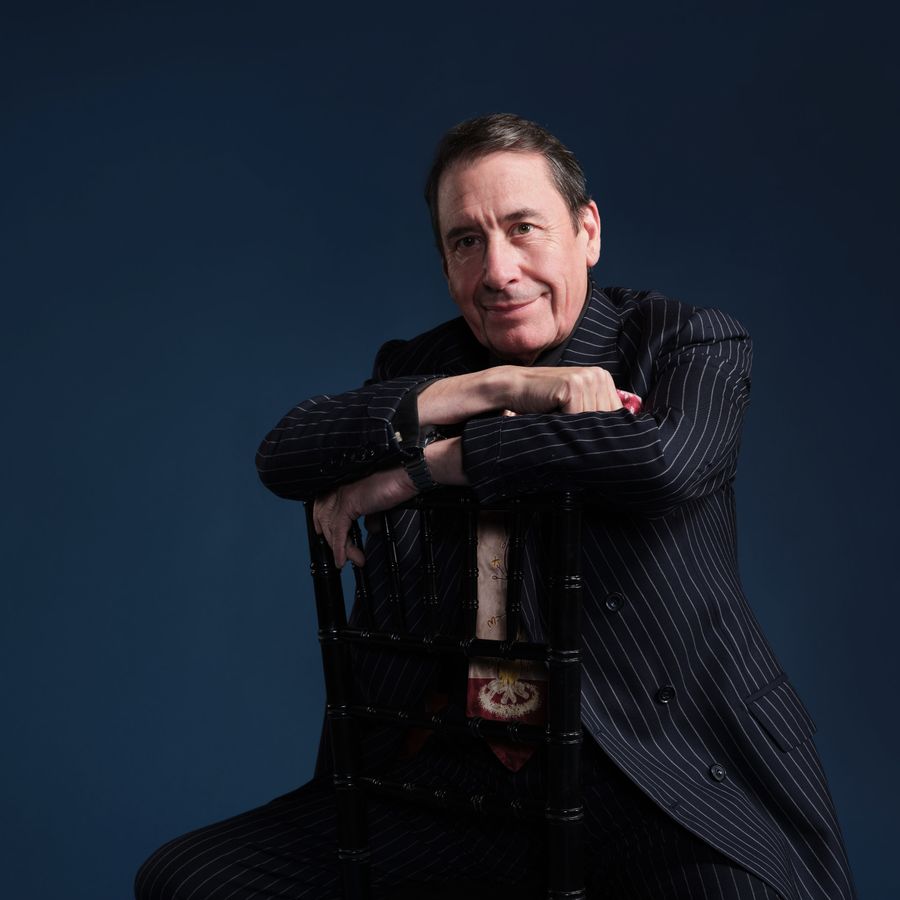 Jools Holland in a suit, casually seated on a chair, looking relaxed and thoughtful