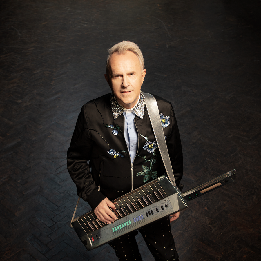Howard Jones, dressed in a suit, stands with a keyboard in hand, looking ready to play