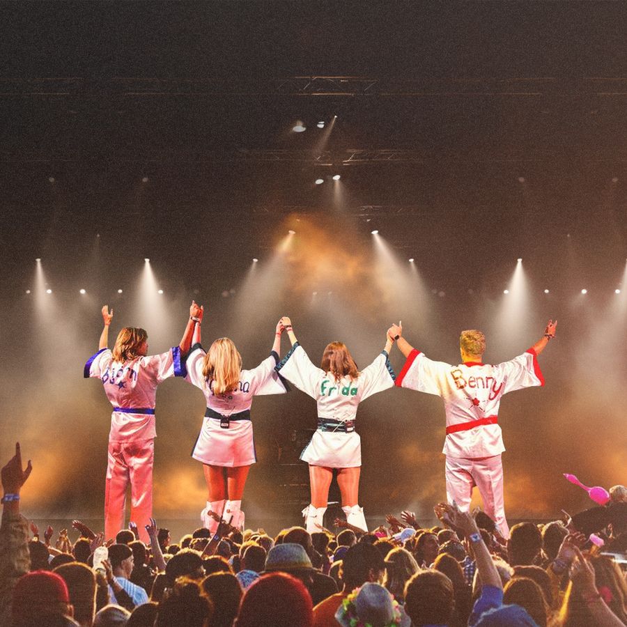 A tribute band, Björn Again, performs on stage with their hands raised, celebrating ABBA's music